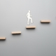 A grey background. There are 6 floating light wood shelves and a figure of a man using the shelves as stairs.