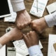 A closeup of four people giving each other a fist bump together over a table covered in office supplies and spreadsheets.