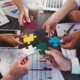 A messy meeting table with reports and coffee cups, etc. There are Multiple arms with hands all meeting in the middle. Each hand holds a different coloured jigsaw puzzle piece.