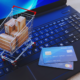 A tiny shopping cart with boxes in it, and two credit cards on the keyboard of a laptop. The screen is blue.