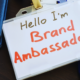 A lanyard with a badge that says "Hello, I'm a Brand Ambassador" on it. There are some notebooks and a laptop keyboard behind it.